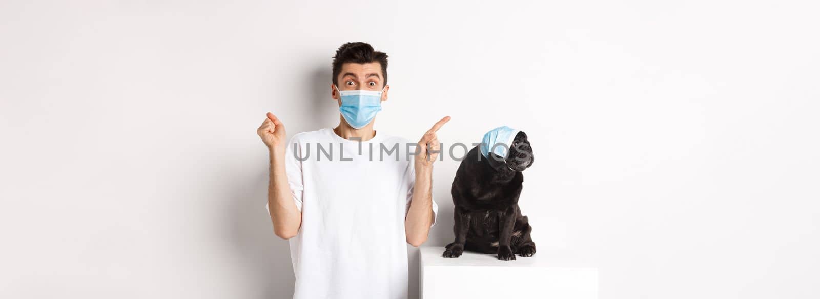 Covid-19, animals and quarantine concept. Young man and black dog wearing medical masks, pug looking at upper left corner and owner showing promo offer, rejoicing by Benzoix