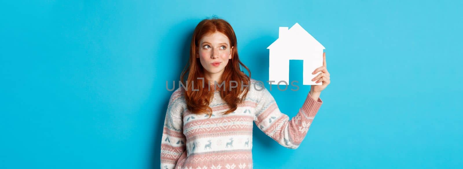 Real estate concept. Image of thoughtful redhead girl showing paper house model and thinking, searching for home or flat, standing against blue background by Benzoix