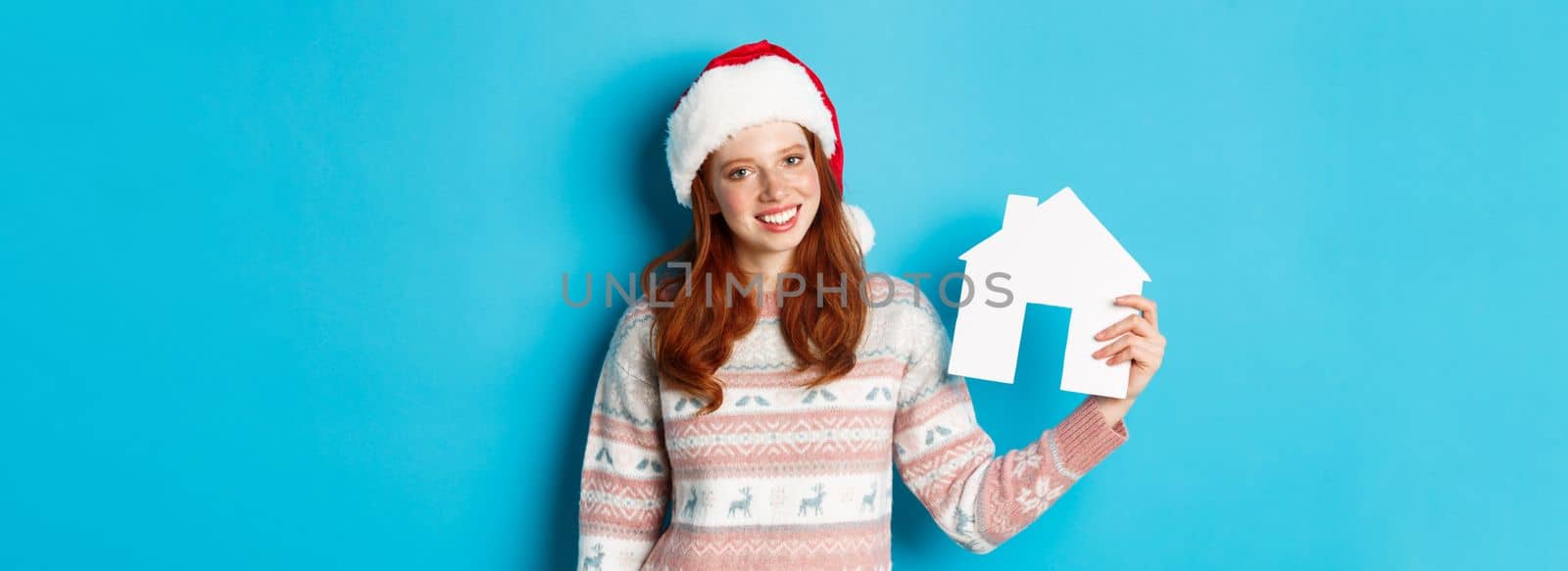 Holiday promos and real estate concept. Cheerful redhead woman in santa hat holding paper house in hand and smiling, standing in sweater against blue background.