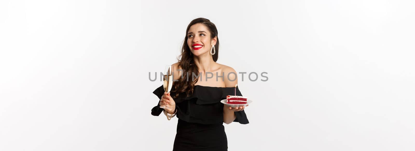 Celebration and party concept. Fashionable woman holding birthday cake with candle and drinking champagne, smiling and looking aside, standing over white background.