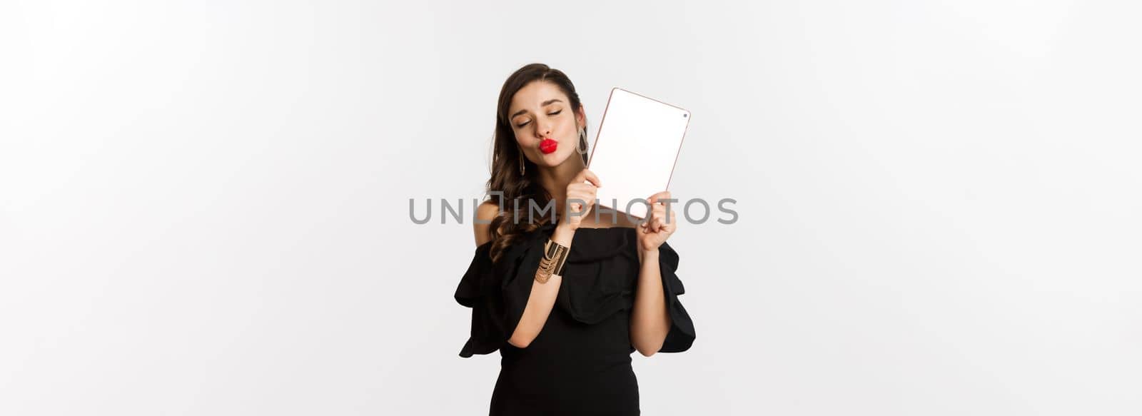 Fashion and shopping concept. Beautiful woman with red lips, wearing black dress, showing digital tablet and making kissing face, white background by Benzoix