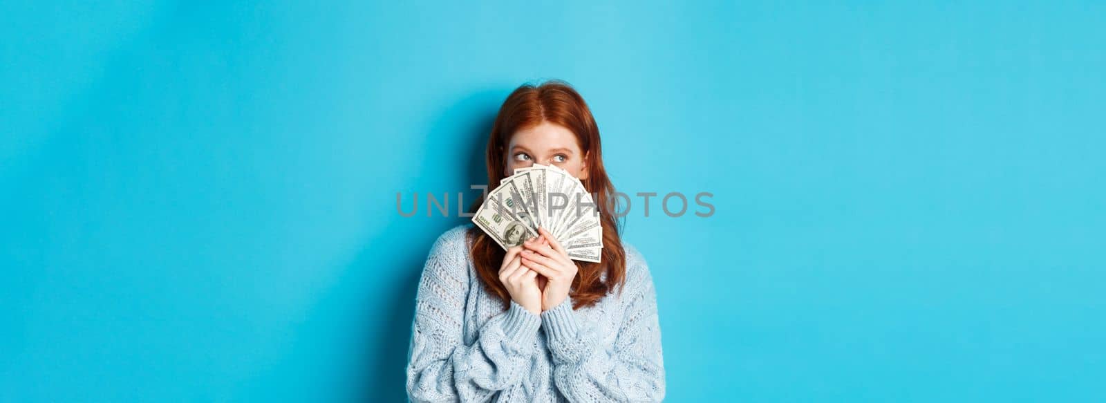 Thoughtful cute girl with red hair dreaming about shopping, holding dollars and looking at upper left corner logo, standing over blue background.