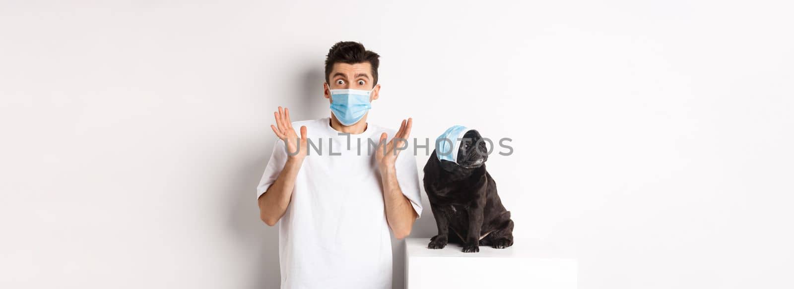 Covid-19, animals and quarantine concept. Shocked dog owner and pug wearing medical masks, staring at camera amazed, standing over white background.