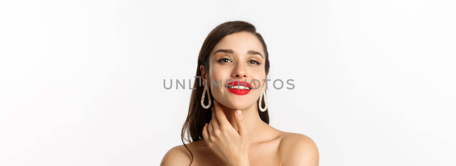 Fashion and beauty concept. Close-up of gorgeous brunette woman with red lips, touching face and smiling self-assured, standing over white background.