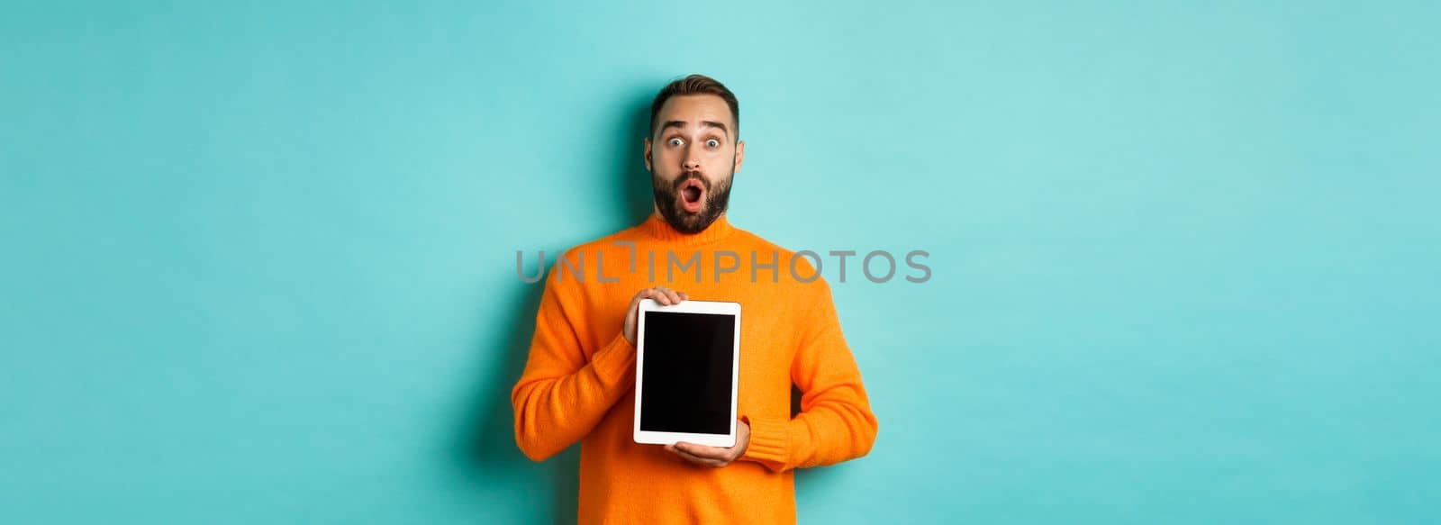 Technology. Surprised bearded man drop jaw, staring at camera and showing digital tablet screen, standing againt light blue background.