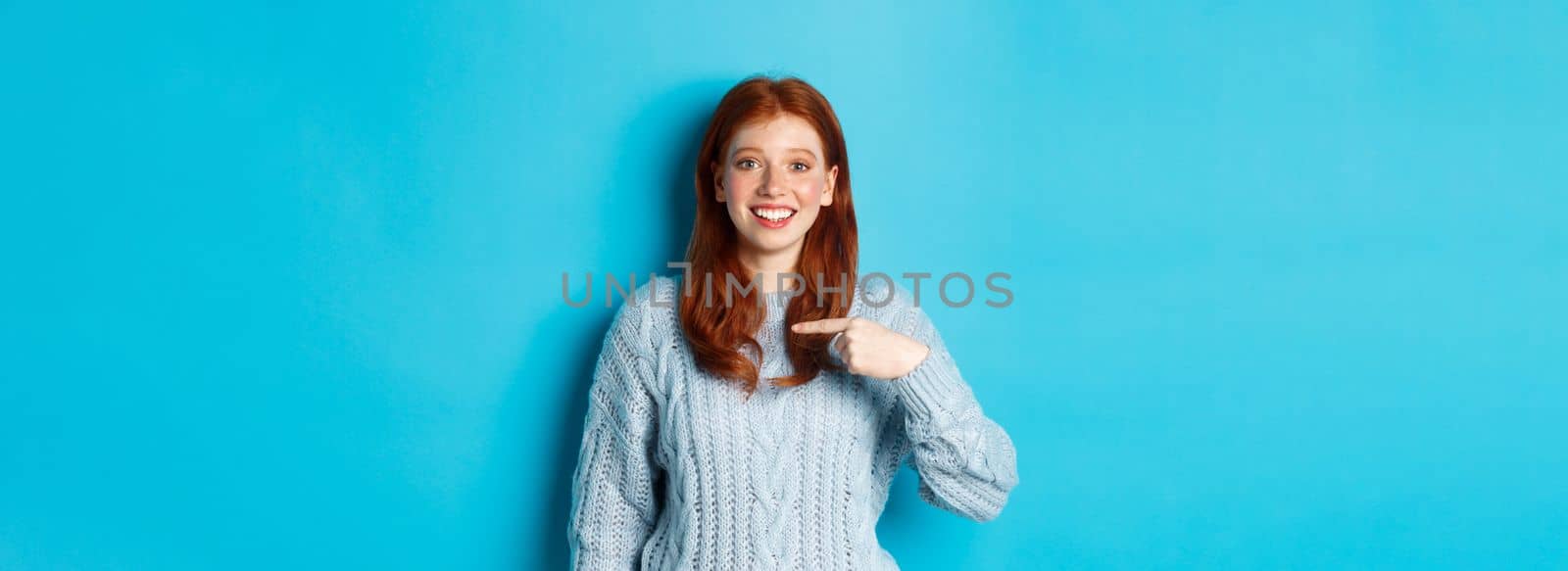 Beautiful redhead girl pointing at herself and smiling happy, being chosen, standing in sweater against blue background by Benzoix