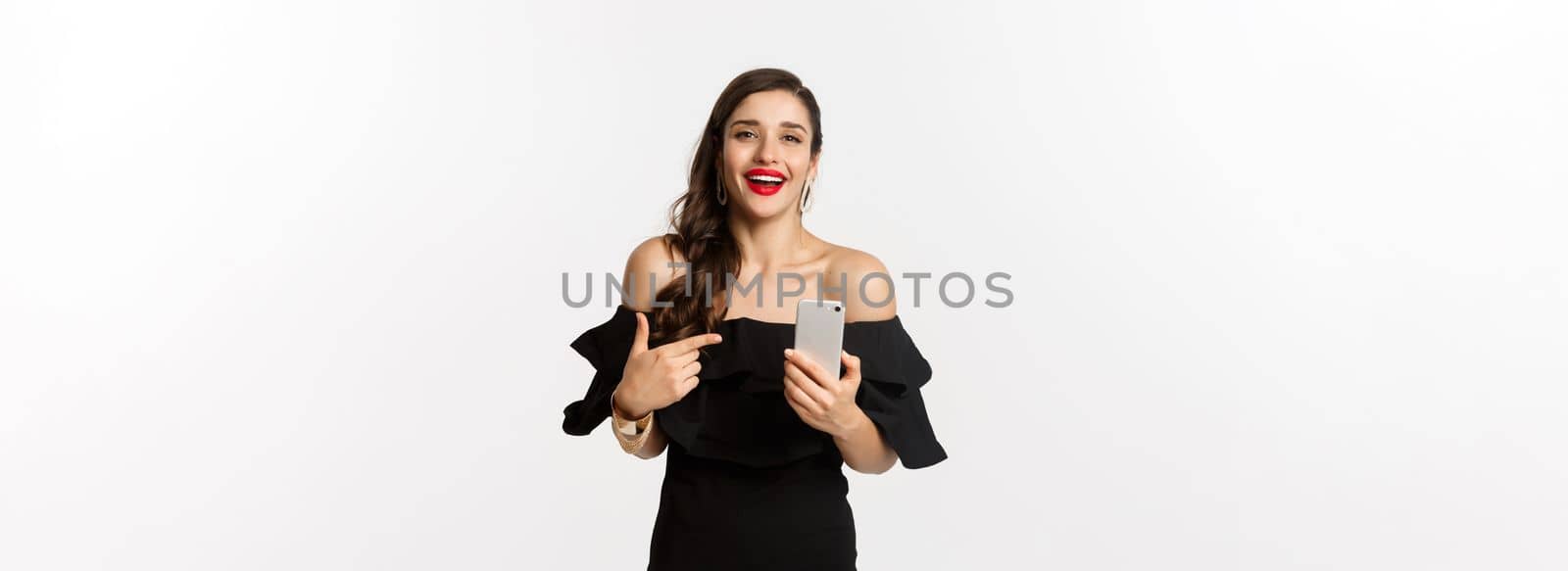 Online shopping concept. Satisfied pretty woman in black dress, smiling pleased and pointing at mobile phone, standing over white background.