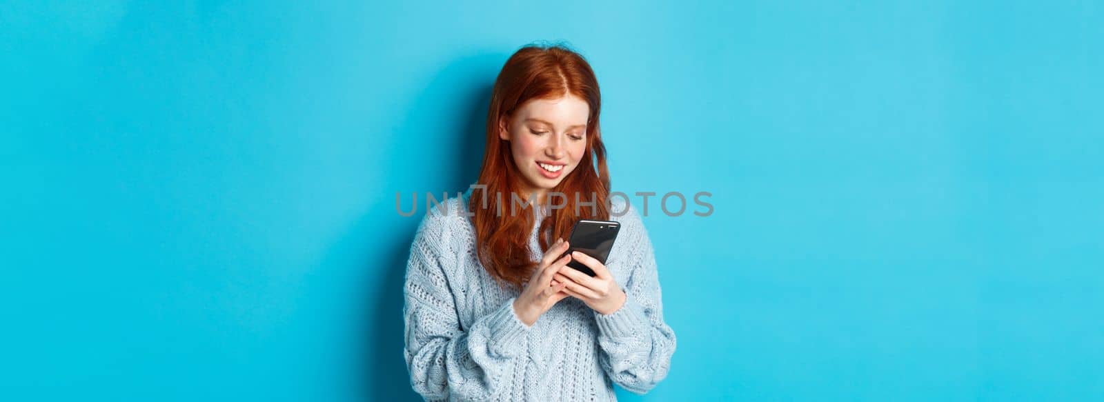 Young teenage redhead girl reading message on smartphone and smiling, using mobile phone and standing over blue background by Benzoix