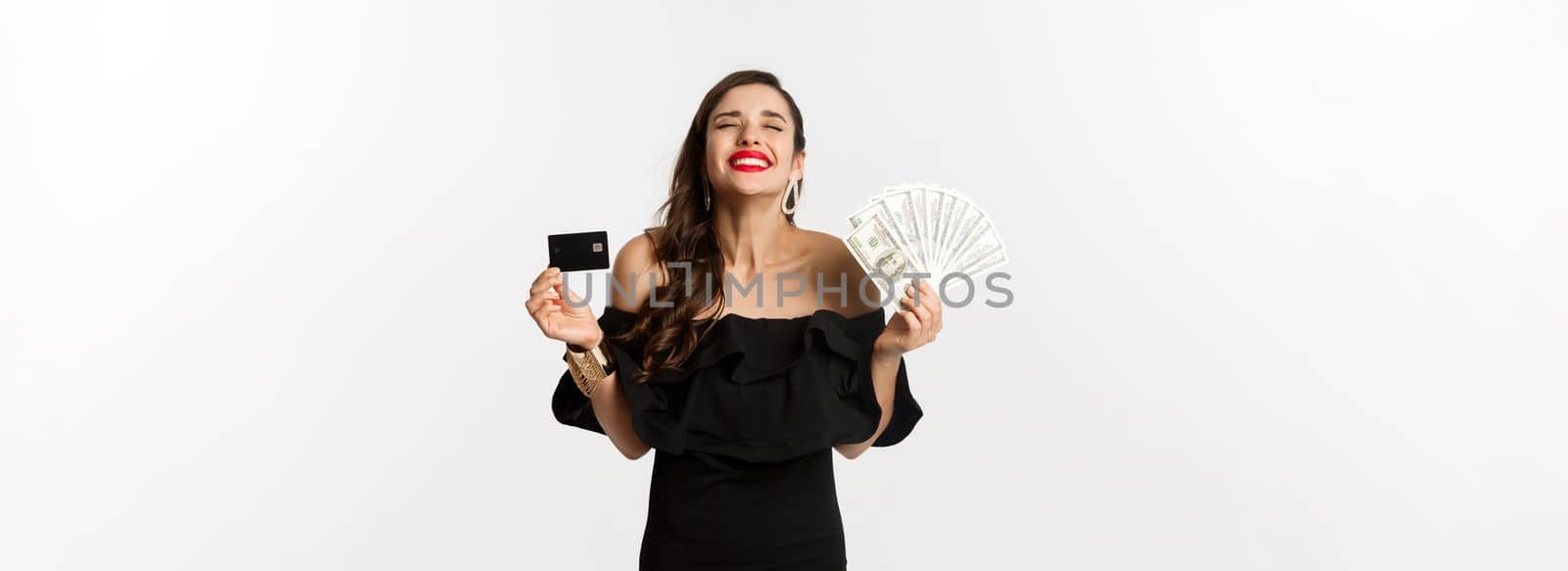 Beauty and shopping concept. Satisfied young woman in stylish dress, looking pleased, holding credit card and money, standing over white background by Benzoix