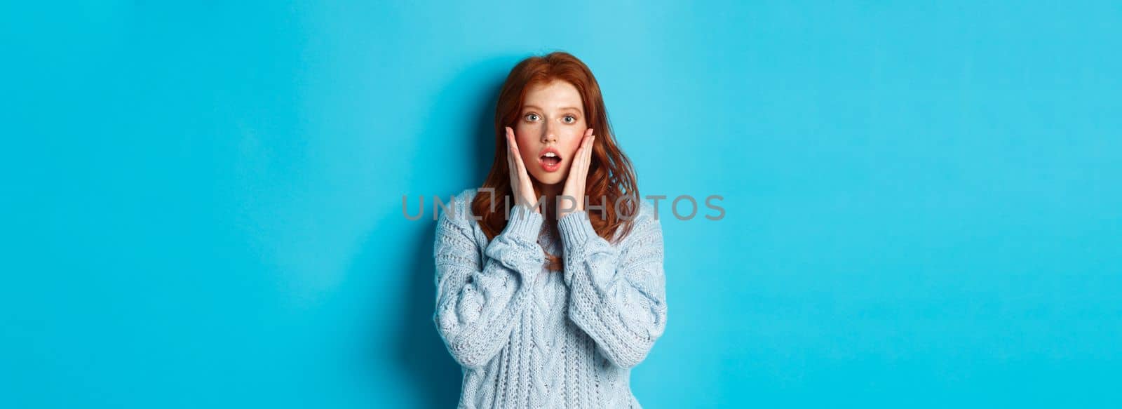 Image of young beautiful redhead girl, looking surprised, staring with complete disbelief at camera, standing in sweater against blue background by Benzoix