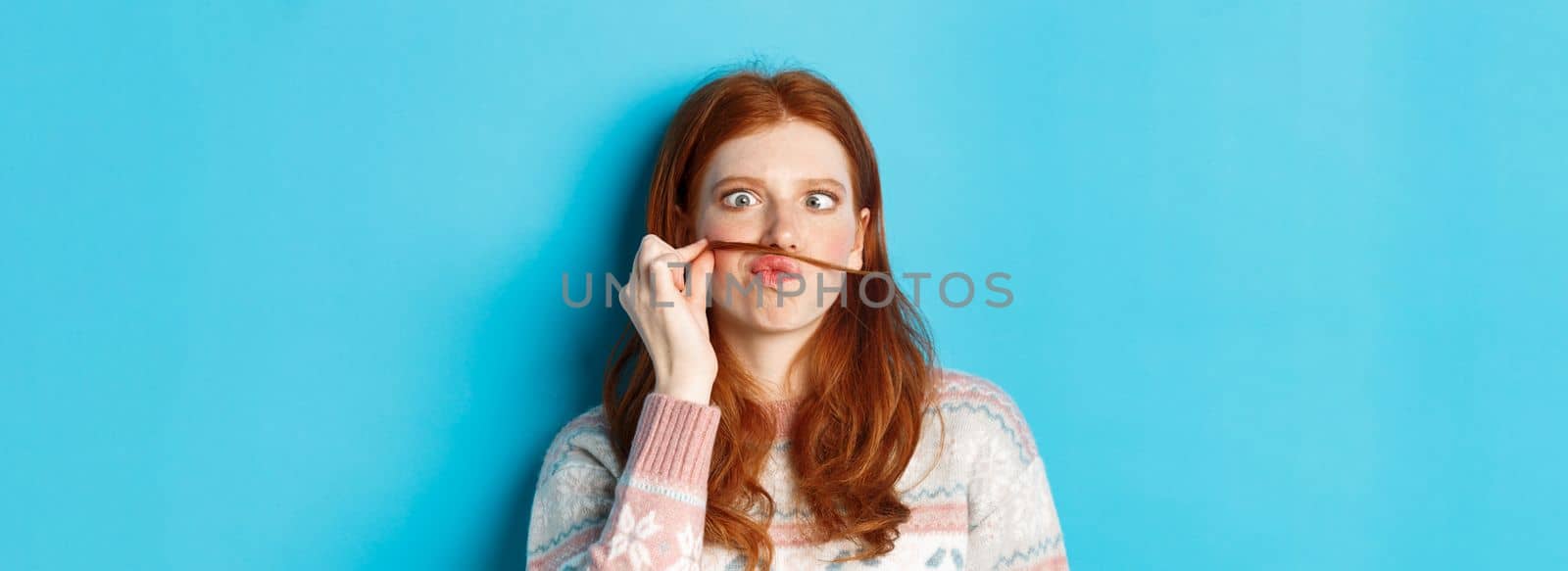 Close-up of silly and funny redhead girl making moustache with hair strand and puckered lips, grimacing against blue background.