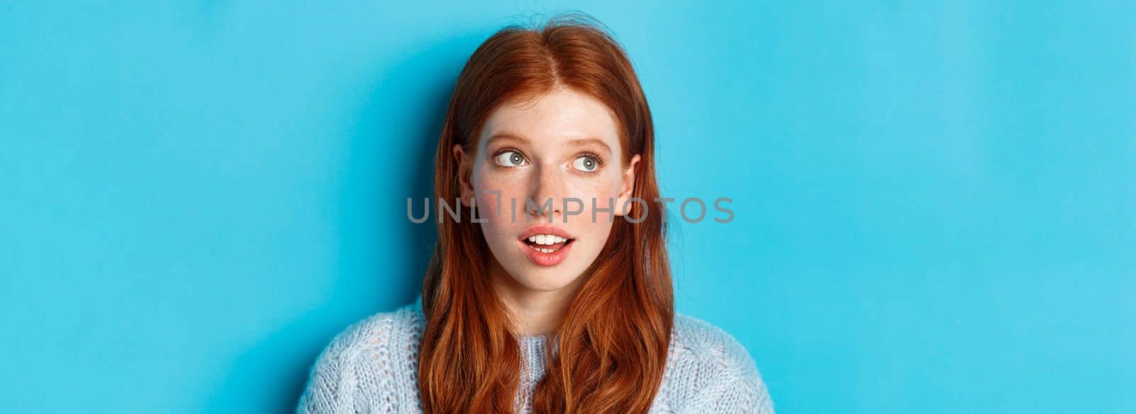 Headshot of thoughtful redhead teen girl looking at upper left corner, staring at logo with curious expression, standing over blue background.