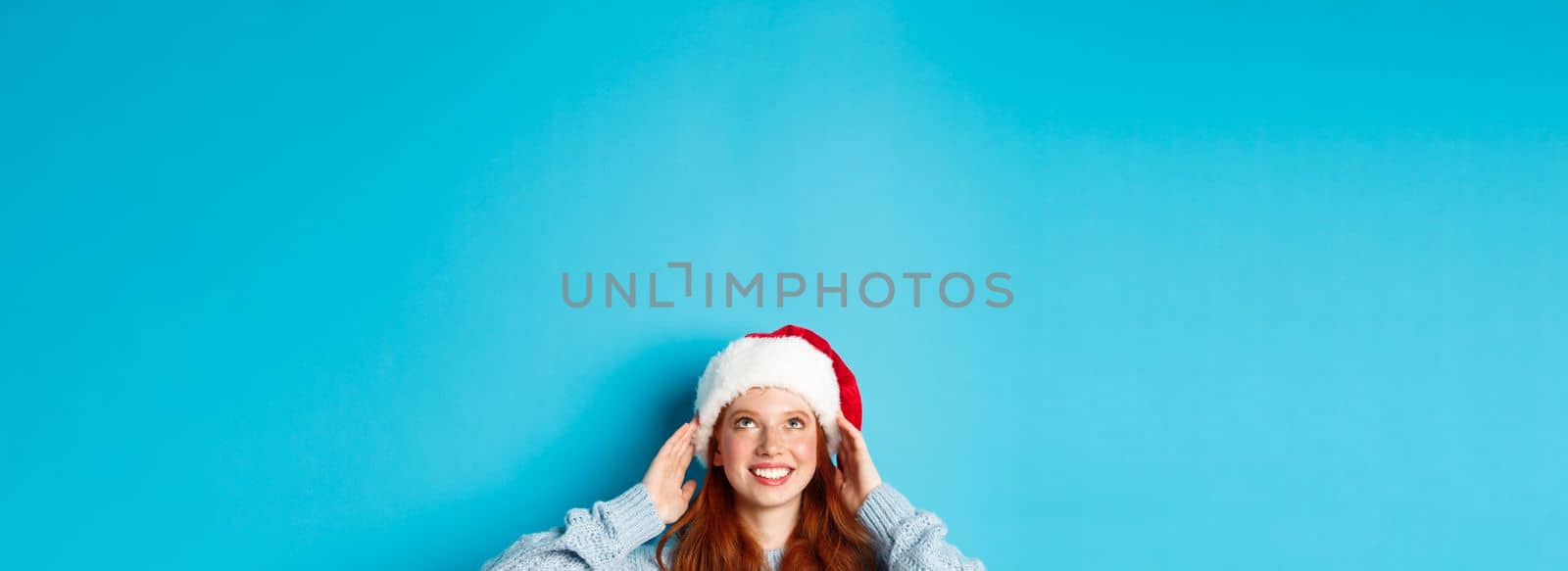 Winter holidays and Christmas eve concept. Head of cute redhead girl in santa hat, appear from bottom and looking up at copy space, staring logo, standing over blue background by Benzoix