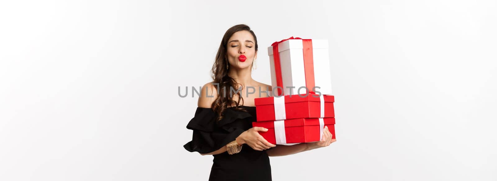 Celebration and christmas holidays concept. Silly woman in elegant black dress, holding xmas and new year presents, pucker lips for kiss, standing happy over white background.