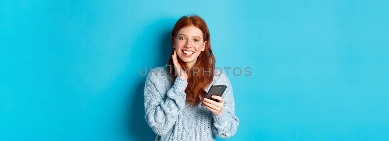 Beautiful redhead girl in sweater, smiling at camera, using mobile phone app, standing with smartphone against blue background by Benzoix