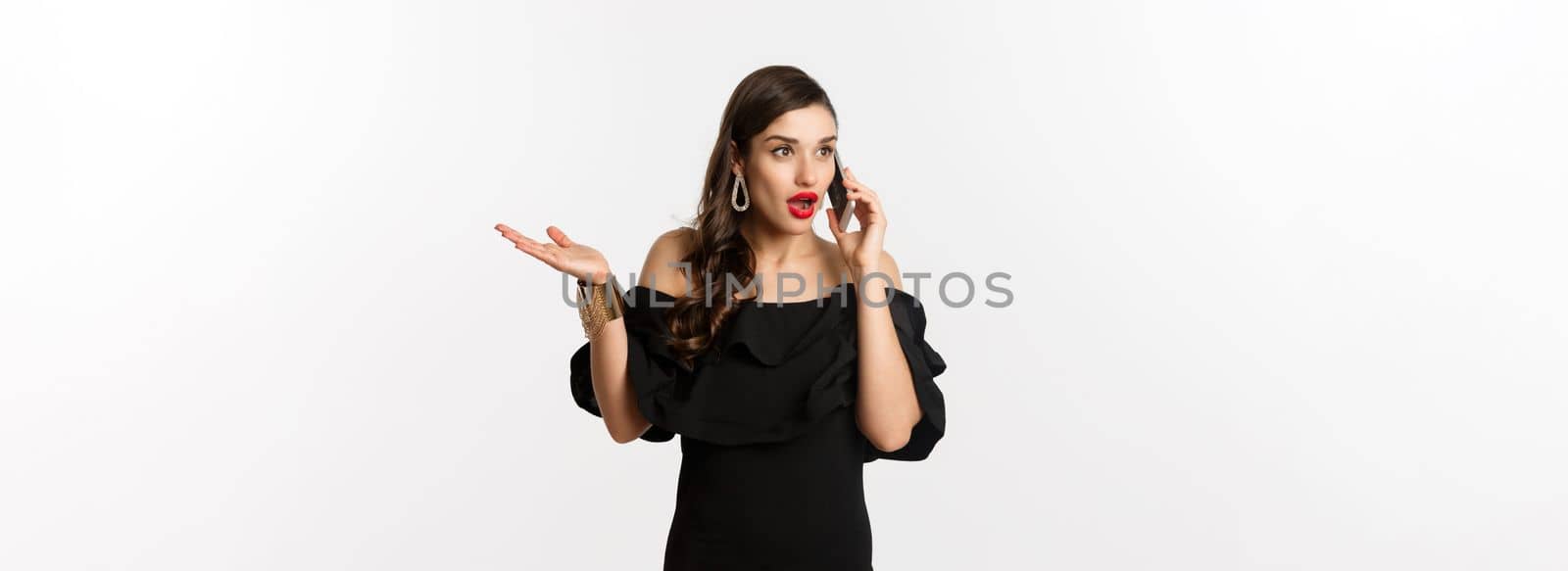 Attractive glamour woman in black dress talking on mobile phone, having conversation and looking surprised, standing over white background.