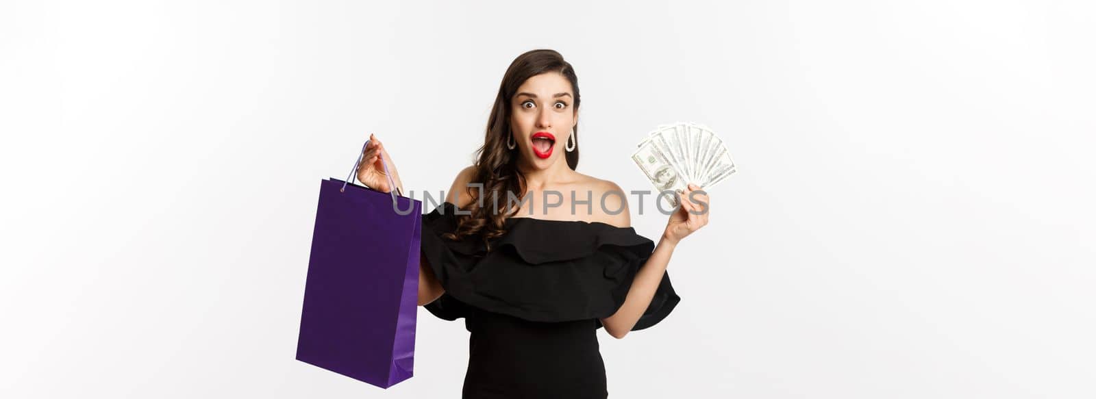 Happy woman buyer holding shopping bag and money, standing in black dress over white background. Copy space