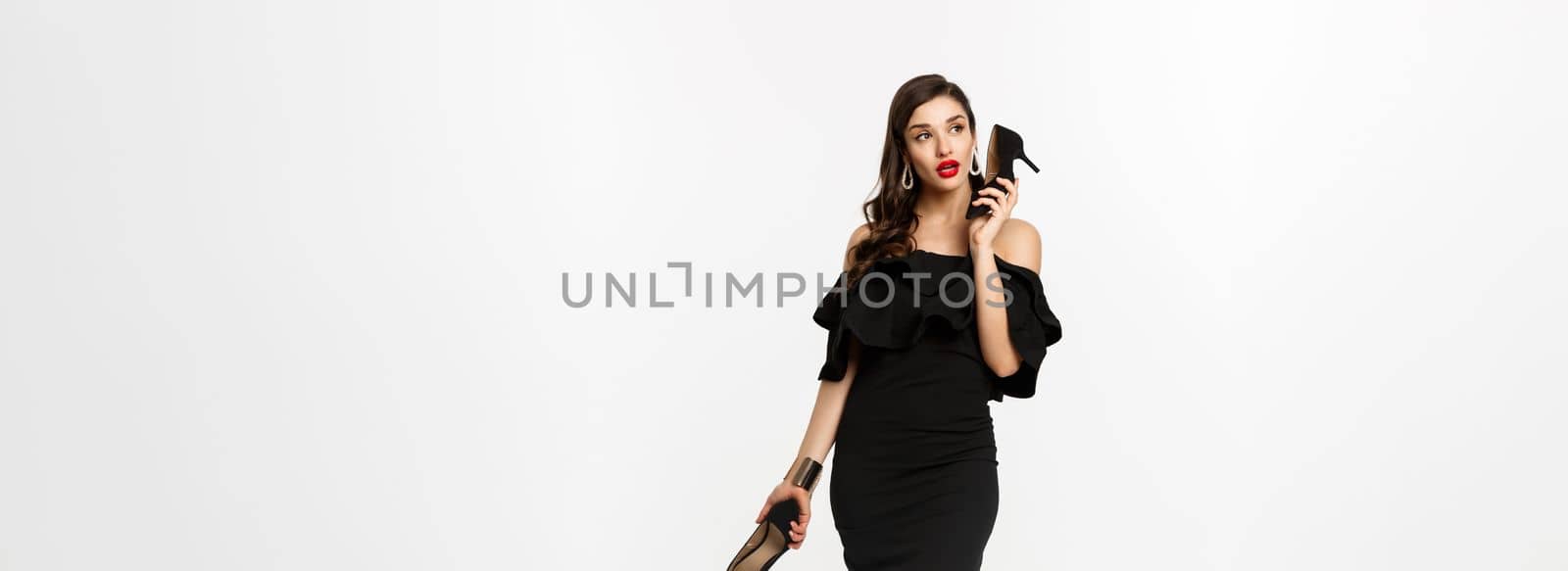 Beauty and fashion concept. Full length of attractive young woman using high heels like mobile phone, standing in black dress against white background by Benzoix