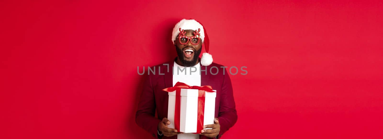 Christmas. Handsome african american man in party glasses and santa hat holding new year gift, looking surprised, receive present and smiling, standing over red background.