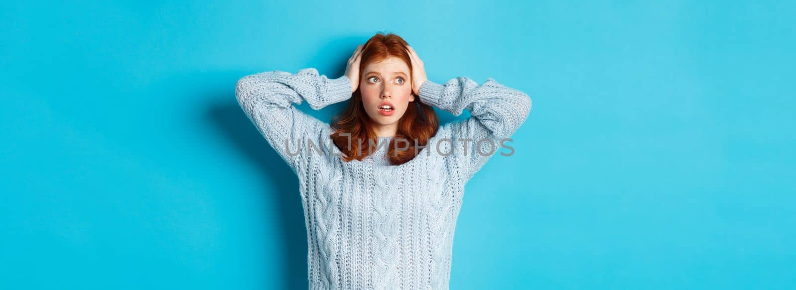 Worried redhead girl standing overwhelmed, holding hands on head in panic and staring left at logo, standing anxious against blue background.