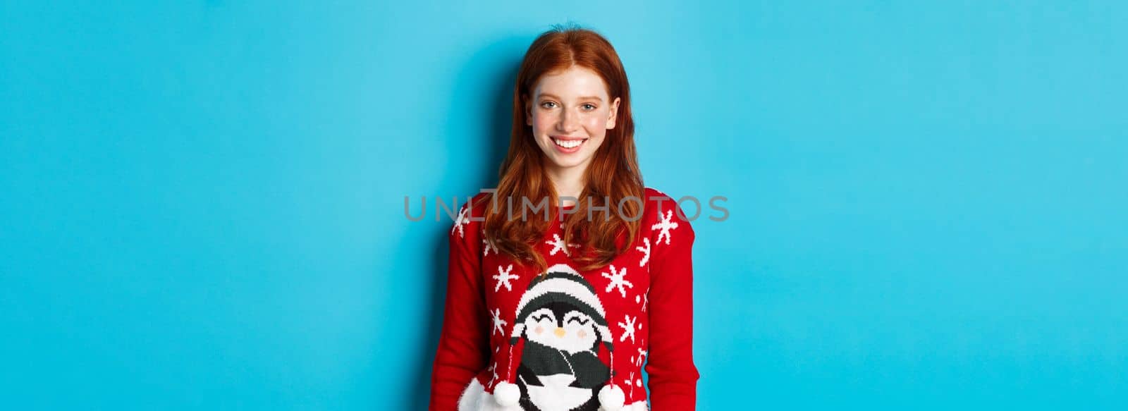 Winter holidays and Christmas Eve concept. Cute smiling teenage girl with red hair, wearing funny xmas sweater, standing against blue background by Benzoix