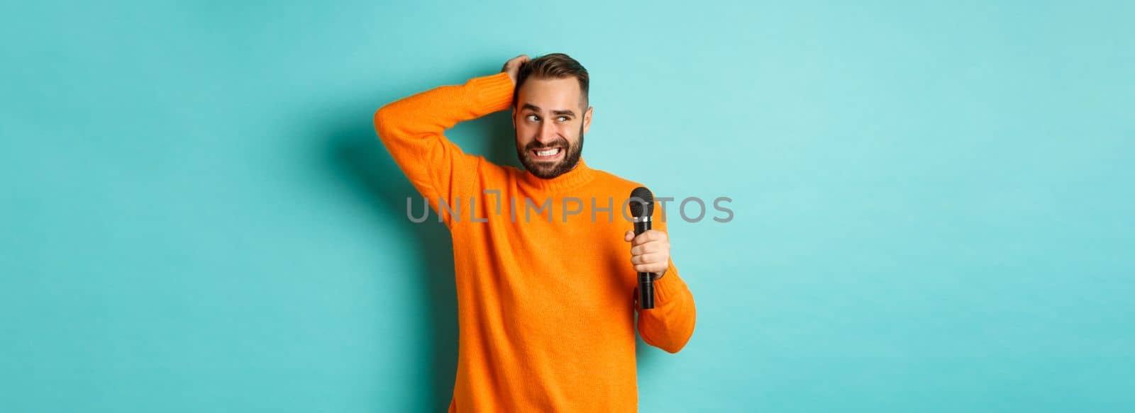 Shy and awkward adult man scratching head, holding microphone before singing, standing over blue background.