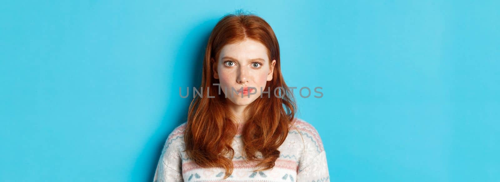 Close-up of serious-looking girl thinking, staring at camera troubled and grimacing, making hard choice, standing over blue background by Benzoix