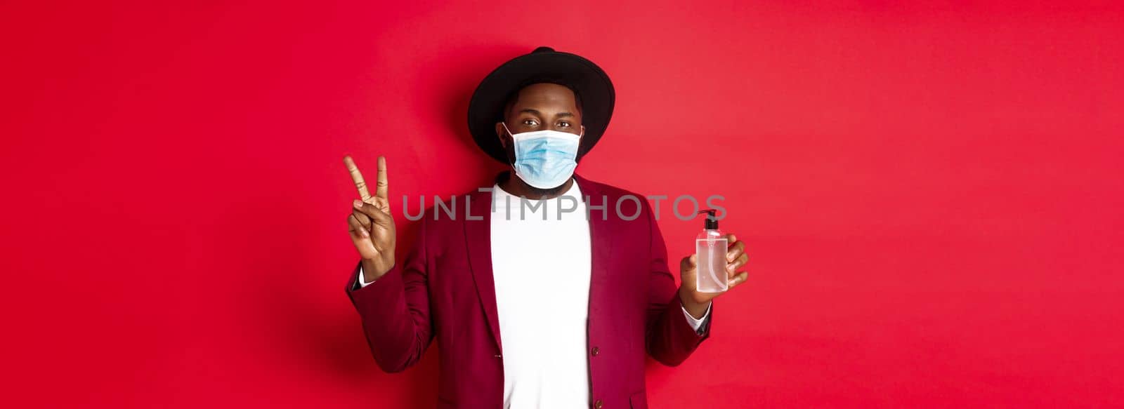 Covid-19, quarantine and holidays concept. Young african american man in medical mask showing hand sanitizer and peace sign, recommending to use antiseptic by Benzoix