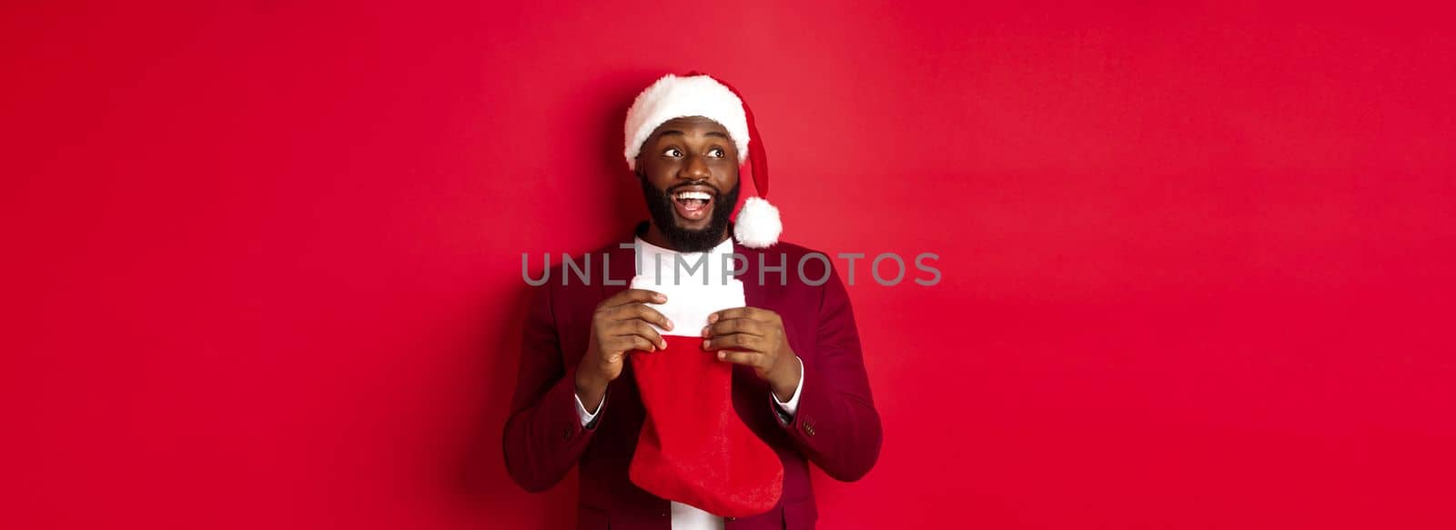 Cheerful Black man looking upper left corner and smiling, holding Christmas sock with gifts, standing over red background by Benzoix