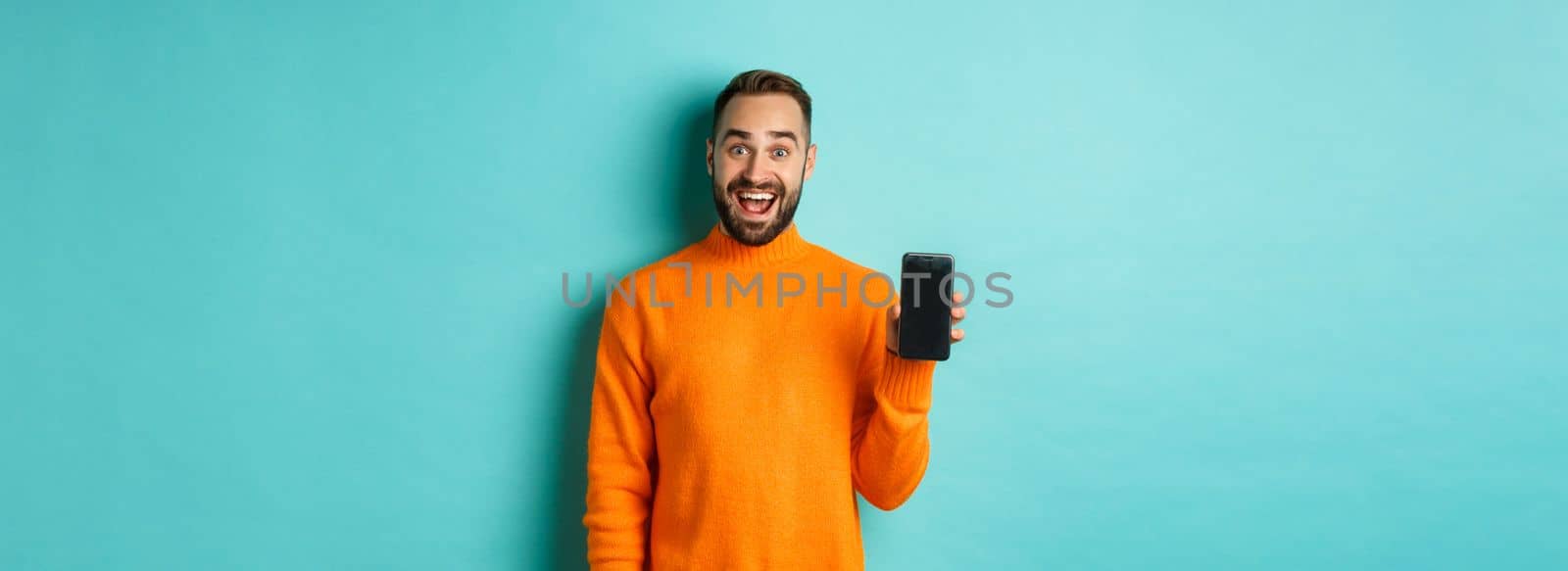 Photo of happy man showing mobile screen, introduce online store, application, standing over turquoise background.
