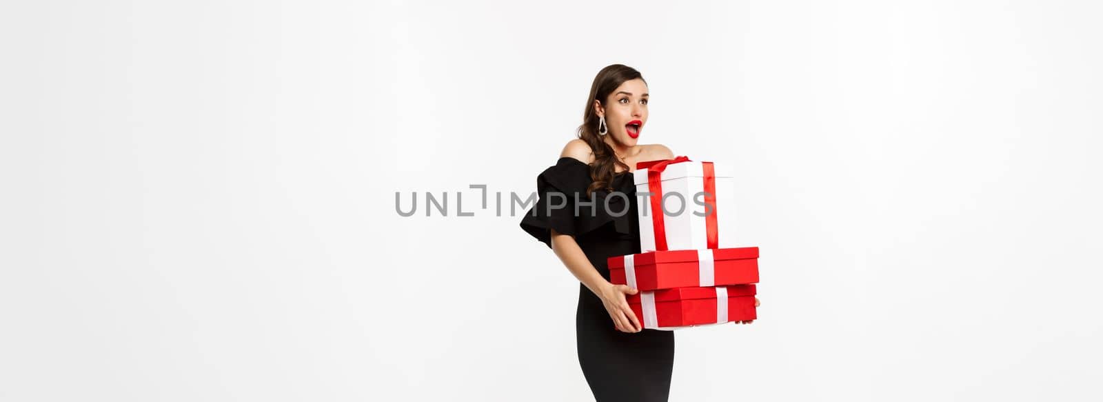 Merry christmas and new year holidays concept. Excited and happy woman in black dress holding xmas presents, looking surprised at logo. standing with presents against white background.