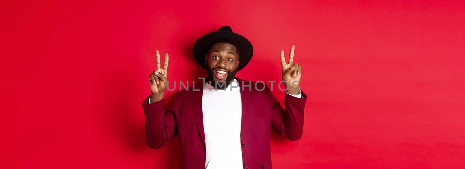 Fashion and party concept. Handsome Black man having fun, showing peace signs and smiling, standing in hat against red background.