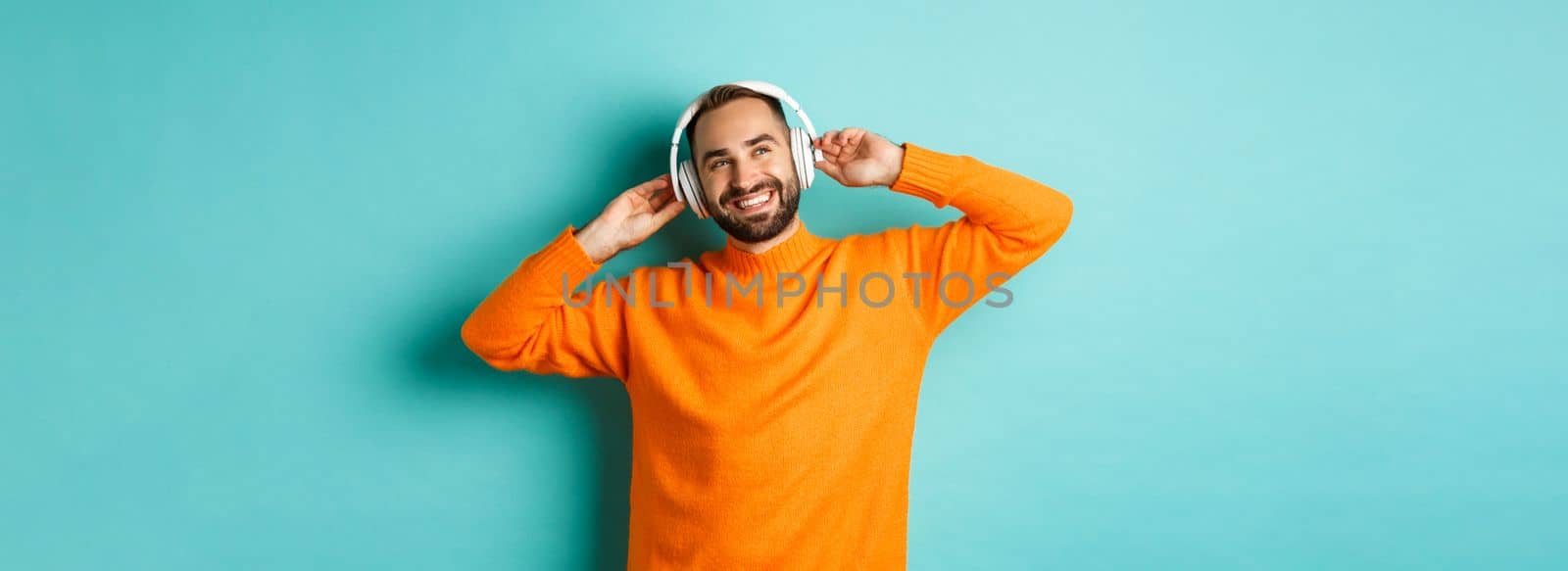 Handsome bearded man listening to music in headphones, smiling pleased, standing over turquoise background by Benzoix