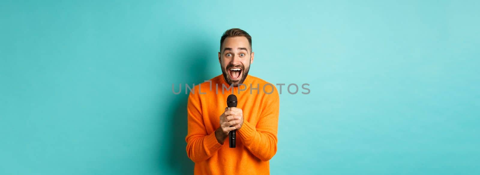 Handsome adult man perform song, singing into microphone, standing against turquoise background.