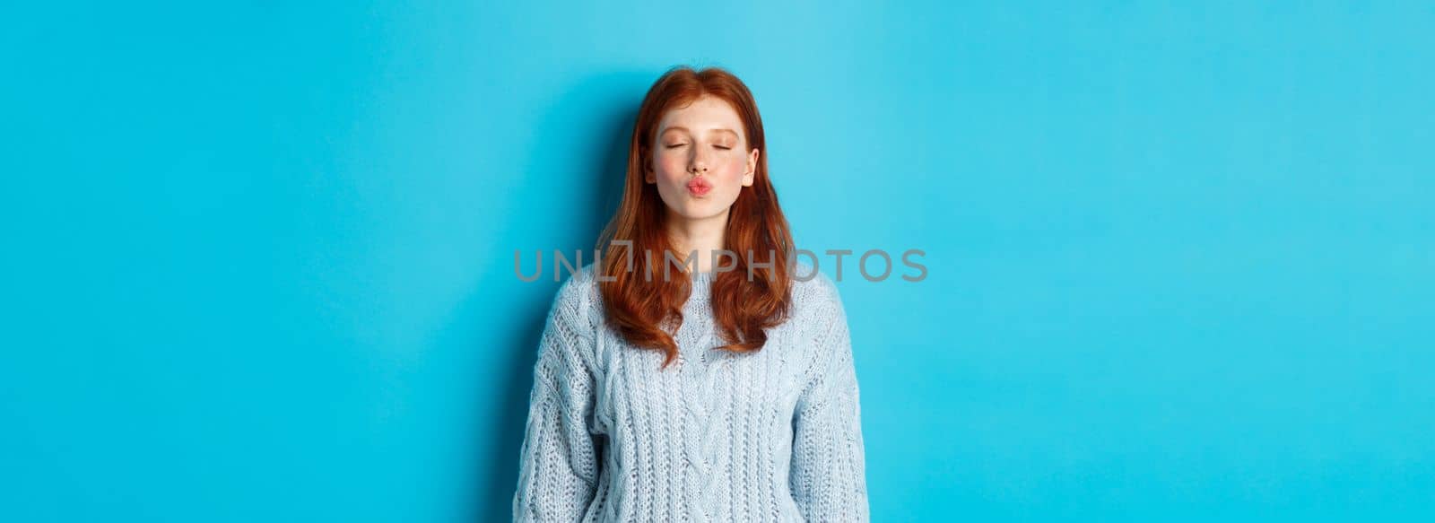 Cute redhead teen girl waiting for kiss, pucker lips and close eyes, standing in sweater against blue background.
