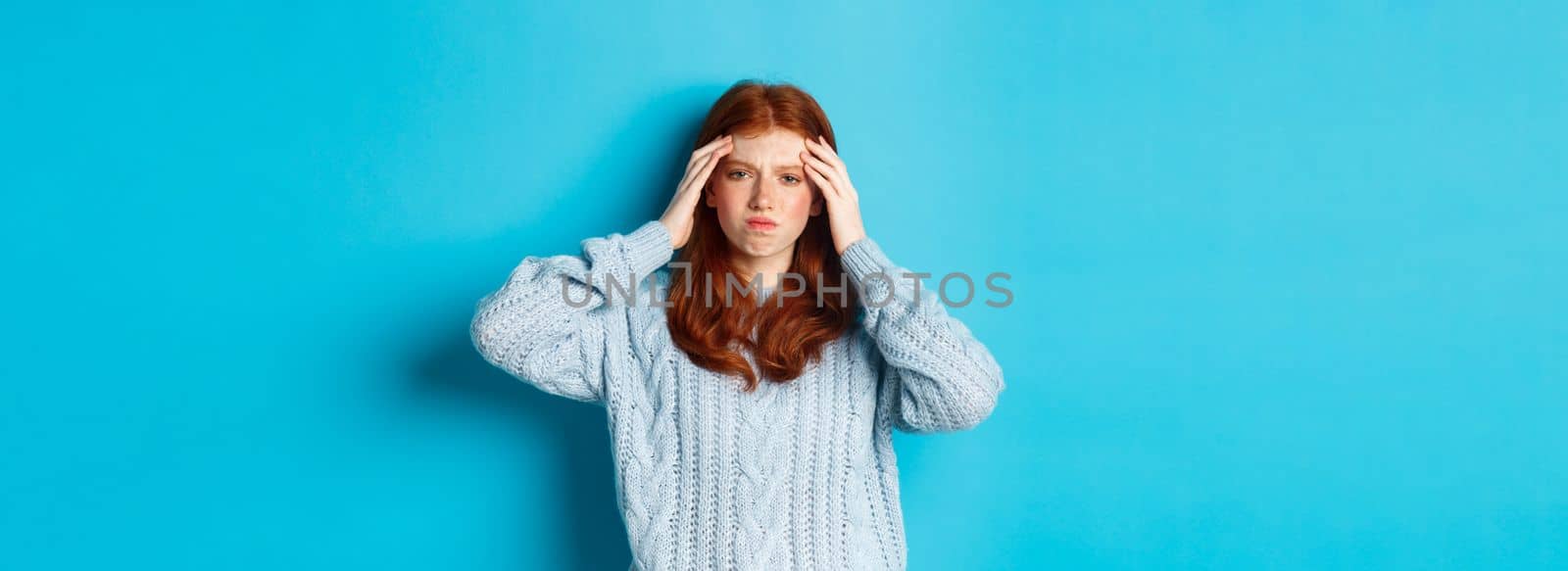 Distressed teenage redhead girl touching head, looking with troubled face expression, standing against blue background, have problem by Benzoix