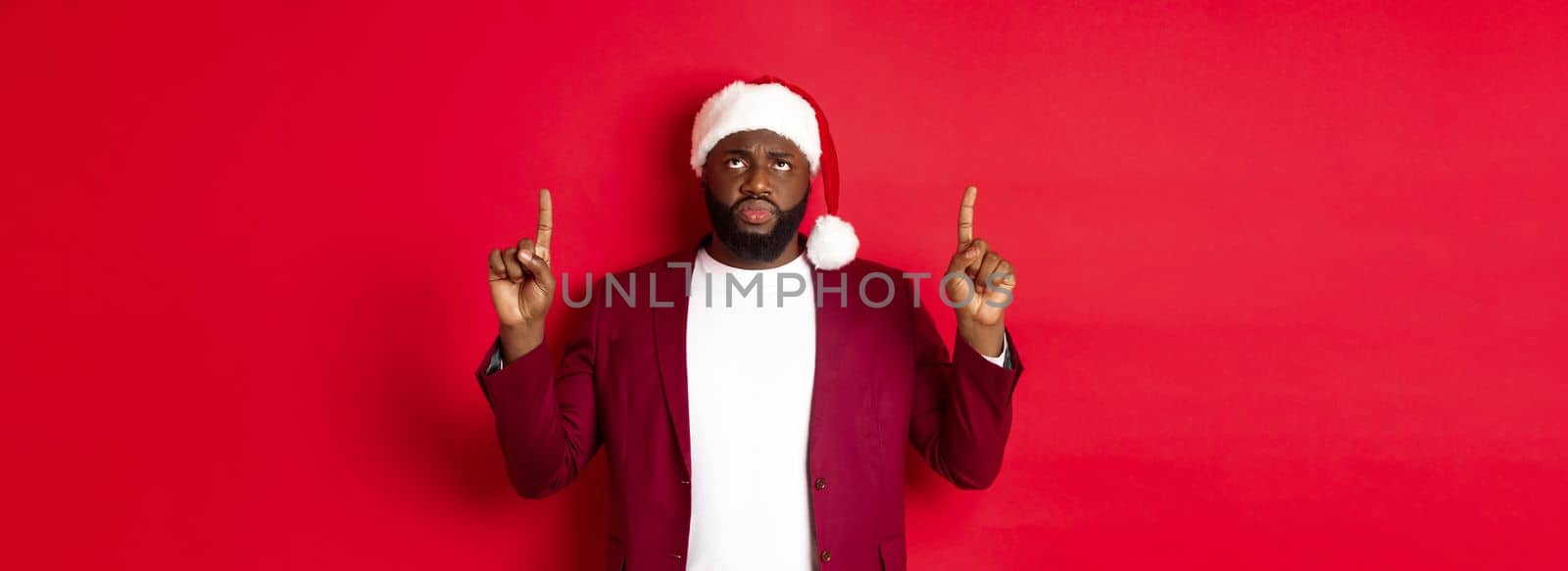 Christmas, party and holidays concept. Upset african-american guy in santa hat complaining, pointing fingers up and frowning disappointed, standing against red background.