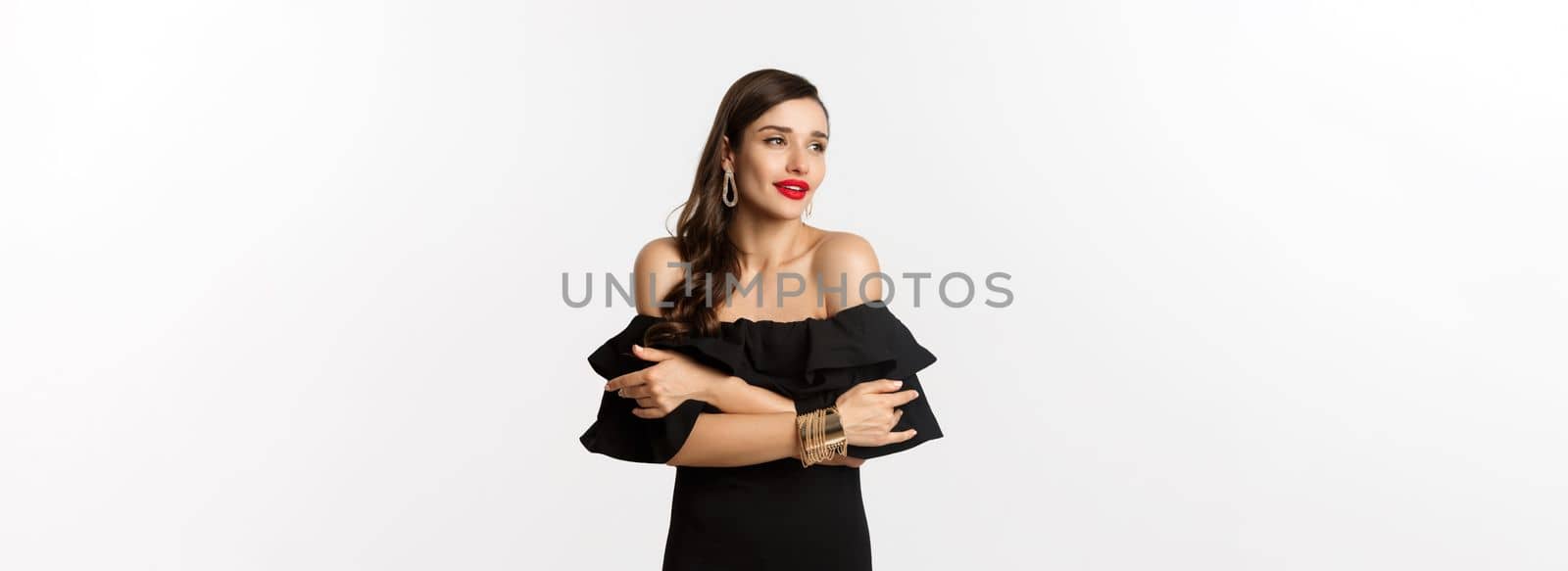 Beauty and fashion concept. Elegant and beautiful woman in black dress, makeup, embracing herself and looking away with sensual gaze, standing over white background.