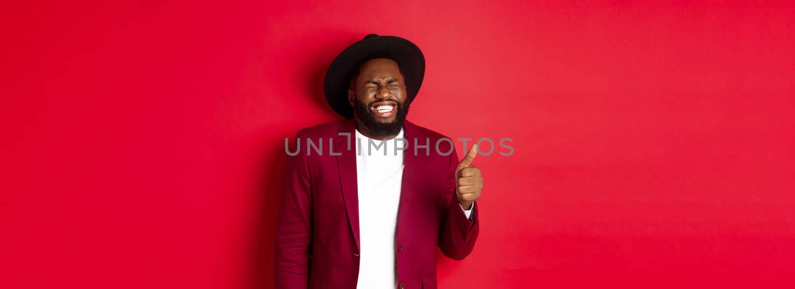 Christmas shopping and people concept. Handsome bearded Black man in party blazer showing thumb up, laughing and having fun, standing over red background by Benzoix