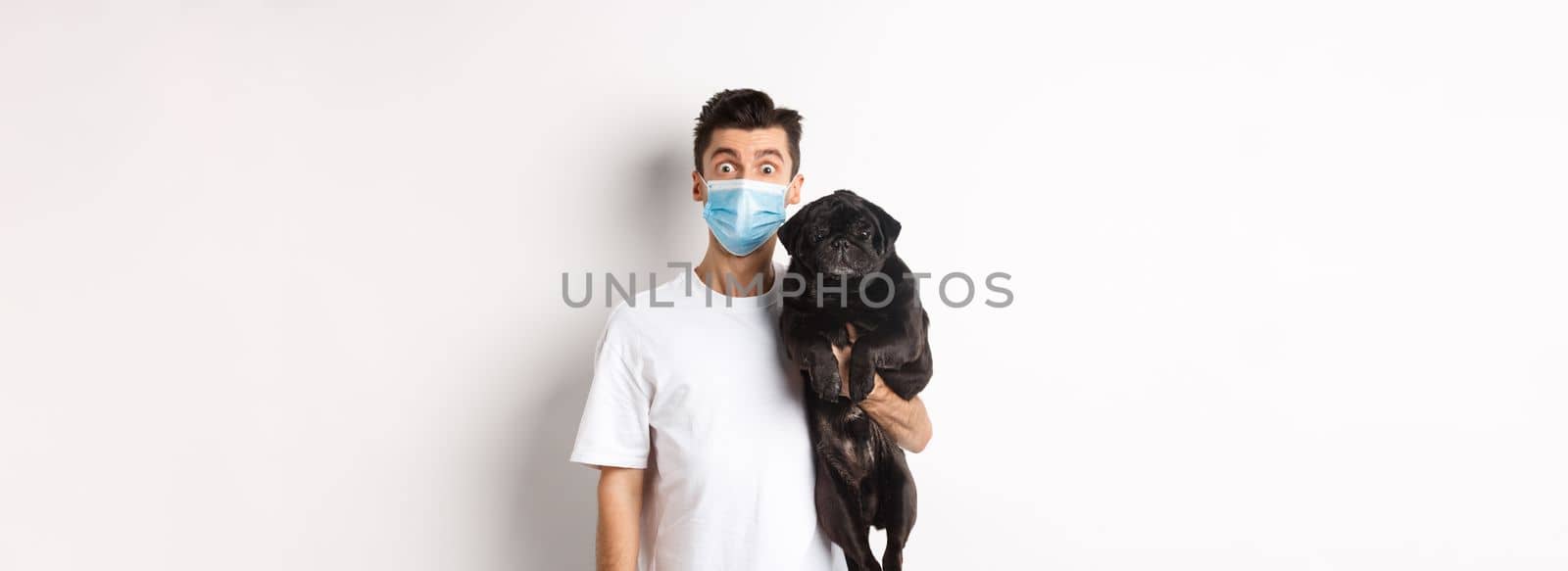 Covid-19, animals and quarantine concept. Funny young man in medical mask holding cute black pug dog, standing over white background.