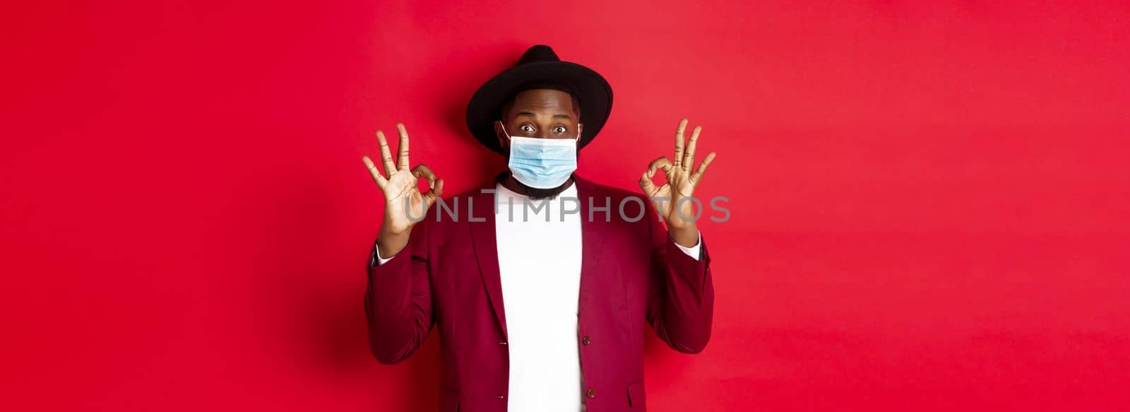 Covid-19 and fashion concept. Stylish african american man in hat and blazer, wearing face mask and showing okay sign, standing over red background.