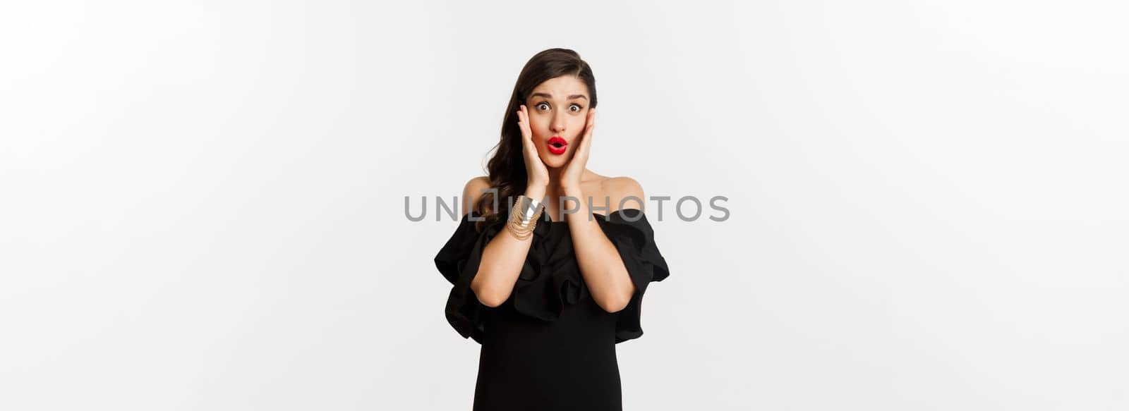 Beauty and fashion concept. Attractive woman in glamour black dress, jewelry and makeup, looking surprised and excited, standing over white background by Benzoix