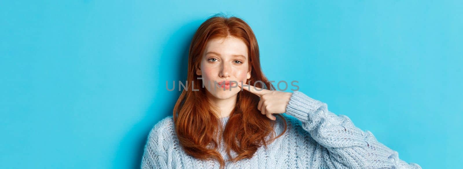 Close-up of sassy teen girl with red hair, pointing at her cheek and staring at camera, standing against blue background by Benzoix