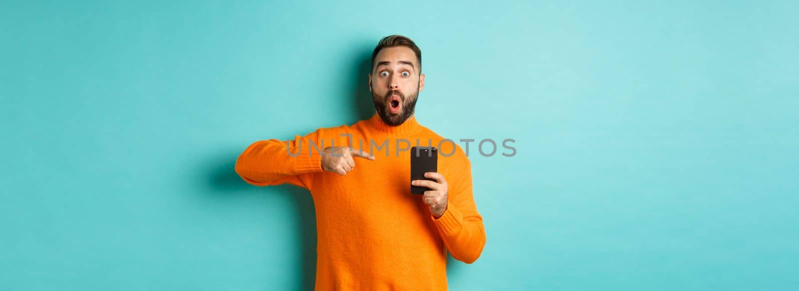Image of impressed man showing something on mobile phone, pointing at smartphone and gasping amazed, standing in orange sweater over light blue background.