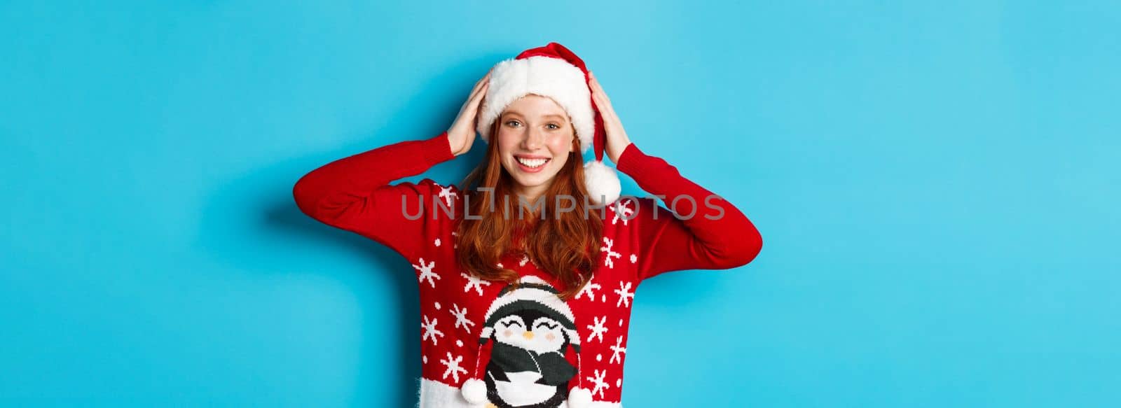 Happy holidays and Christmas concept. Pretty redhead girl in xmas sweater, put on santa hat and smiling, celebrating New Year, blue background.