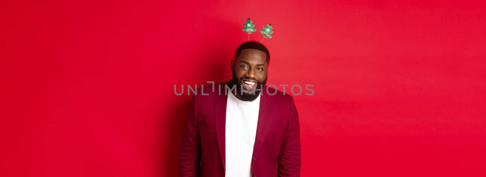 Merry Christmas. Handsome african american man in blazer and party headband, celebrating new year, smiling happy at camera, red background.