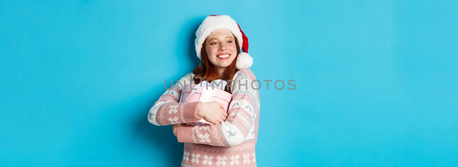 Winter and celebration concept. Dreamy redhead girl in santa hat hugging her christmas gift and looking left, smiling happy, standing over blue background.