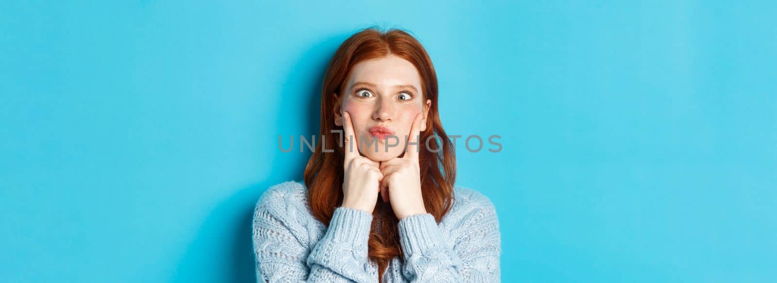 Close-up of funny redhead teen girl making faces, squinting and pocking cheeks, standing against blue background.