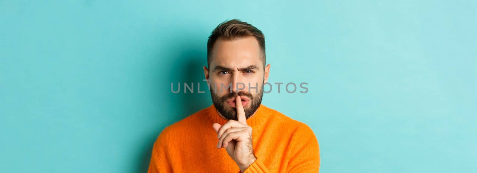 Close-up of annoyed frowning man shushing at camera, press finger to lips, showing taboo gesture, standing over light blue background.