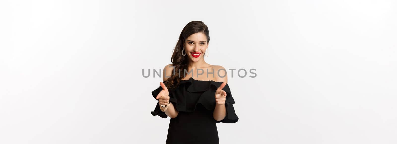 Fashion and beauty. Beautiful young woman in black dress, wearing makeup, pointing fingers at camera to congratulate or praise, smiling pleased, standing over white background by Benzoix